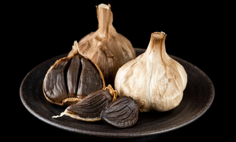 Schwarzer Knoblauch auf einem Teller: aufgeschnittene Knolle mit tiefschwarzen, weichen Zehen neben einer ganzen Knolle.